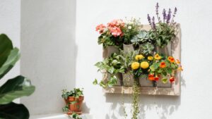 A close-up shot shows a wooden frame holding several fabric pockets filled with various blooming flowers and leafy plants, mounted against a bright wh