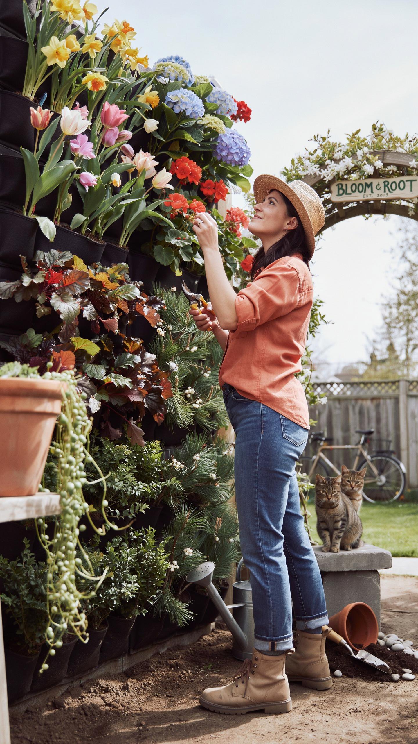 A woman in a straw hat, orange shirt, and jeans is tending to a lush vertical garden filled with colorful flowers, while a cat watches from a low wall